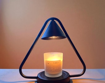 May include: A black candle warmer with a triangular frame and a conical shade. A vanilla-scented candle sits on the base, with a label visible. The warmer is designed to gently melt the candle, releasing its fragrance.