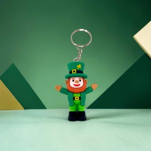 May include: A green leprechaun keychain with a silver keyring. The leprechaun has a red beard, a green hat with a gold shamrock, and a green outfit. The background is a mix of green geometric shapes.