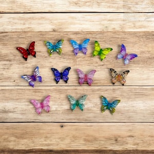 May include: A collection of colorful butterfly decorations in various sizes and designs. The butterflies are in shades of red, blue, purple, green, and pink, with glitter accents. They are arranged on a wooden surface.