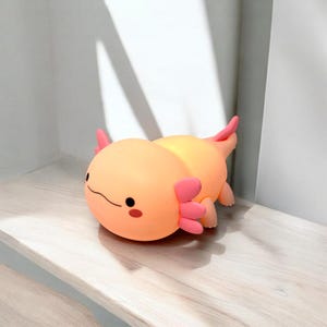 May include: A soft, orange axolotl plush toy with pink gills and accents. The cartoon-style salamander has a smiling face with black eyes and a small mouth. The plush is lying on a light-colored wooden surface.