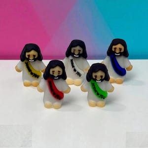 May include: Five small figurines of Jesus with black hair and beards, wearing white robes and colorful sashes. Each sash has the text "Jesus ❤ You" in a different color: yellow, red, white, blue, and green. The background is a gradient of pink and blue.