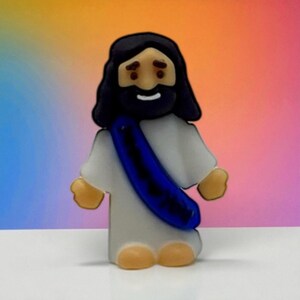May include: A figurine of Jesus Christ, depicted with dark hair and a beard, wearing a white robe and a blue sash. The figure has tan hands and feet, with a smiling expression. The background is a gradient of colors.