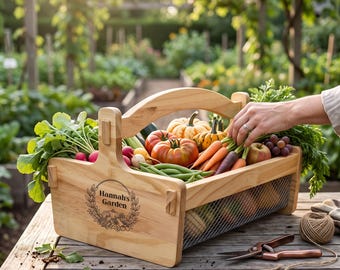 May include: A light-colored wooden harvest basket with a wire mesh base and a central handle. The side is engraved with a wreath design and the text "Hannah's Garden." The basket is filled with fresh produce including tomatoes, carrots, radishes, and small pumpkins.