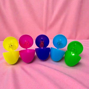 May include: A row of colorful plastic Easter eggs, each split open to reveal a small toy animal inside. The eggs are in shades of yellow, pink, blue, and green, set against a pink background.