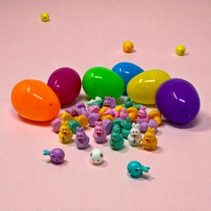 May include: A collection of colorful Easter eggs and miniature figurines on a pink surface. The eggs are in various colors, including orange, purple, green, blue, and yellow. The figurines are small, depicting bunnies and other Easter-themed characters.