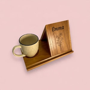 Puede incluir: Un soporte de madera con un panel triangular grabado con el nombre Emma y un dibujo floral, acompañado de una taza de cerámica color crema con motas que descansa sobre una base circular integrada en la bandeja de madera.