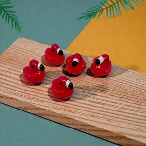 May include: Five small, red flamingo-shaped beads with black and white details are arranged on a wooden surface. The beads have a glossy finish and are set against a backdrop of teal and yellow.