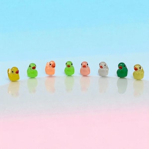 May include: A row of eight miniature duck figurines in various colors, including yellow, green, orange, pink, and white. Each duck has a small orange beak and black eyes. The background is a gradient of blue and pink.