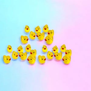 May include: A collection of small, yellow duck figurines with red beaks and black eyes. The ducks are scattered on a gradient background of blue and pink. These miniature ducks are likely decorative items or craft supplies.