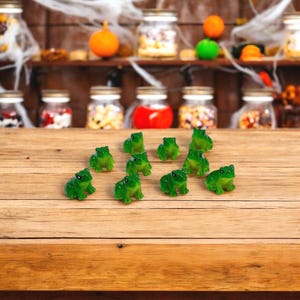 May include: A collection of eleven small, translucent green frog figurines arranged on a wooden surface. Each miniature frog features a smooth, glossy finish with simple, molded details, including defined eyes and a seated posture, creating a uniform set.