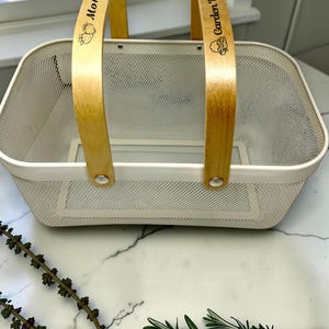 May include: A rectangular white wire mesh basket with two light-colored wooden handles. The left handle is engraved with the text Mom's and strawberry icons, while the right handle features the text Garden Basket with vegetable icons.