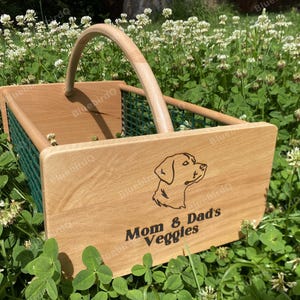 May include: A rectangular wooden harvest basket with a curved handle and green wire mesh sides. The front wooden panel features a laser-engraved outline of a dog head and the text "Mom & Dad's Veggies" in a dark, bold, sans-serif font.
