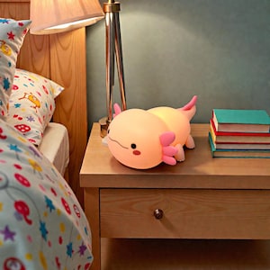 May include: A soft, glowing axolotl night light in shades of peach and pink rests on a wooden nightstand. The lamp has a smiling face and is next to a stack of books. A lamp and bed with a patterned comforter are in the background.