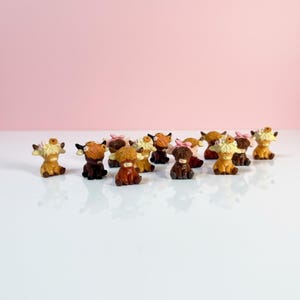 May include: A collection of small, miniature highland cow figurines in various shades of brown, tan, and cream. Each figurine features a shaggy coat, small horns, and some are adorned with tiny floral accents or small bows on their heads.