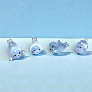 May include: Four small, translucent blue seal figurines with black whiskers and eyes. The seals are in various poses, with flippers and tail details. They are arranged on a light blue surface against a matching background.
