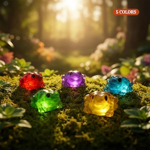 May include: Five small, translucent frog figurines in red, purple, blue, green, and yellow, each featuring dark eyes. The frogs are arranged on a patch of green moss. An orange rectangular label in the upper right corner displays the text 5 COLORS.