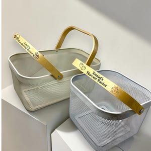 May include: Two rectangular wire mesh baskets with wooden handles. The larger cream-colored basket features the text "Emily's Gardening Basket" with a small icon. The smaller silver-colored basket features the text "Daniel's Harvest Basket" with a berry icon.