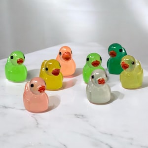 May include: A collection of nine colorful, translucent duck figurines. The ducks are in various shades of green, yellow, pink, orange, and white, each with a red beak. The figurines are arranged on a white surface.