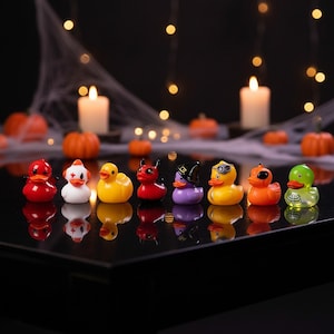May include: A collection of eight small rubber ducks arranged in a row on a reflective black surface. The ducks feature various designs including a red devil, a white ghost, a classic yellow duck, a purple witch, a green skeleton, and orange figures.