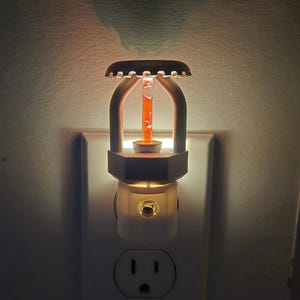 May include: A white night light with a decorative design. The night light has a cylindrical glass tube with orange liquid inside, and a decorative top. The night light is plugged into a wall outlet.