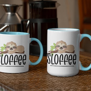 May include: Two white ceramic mugs with a blue rim. The mugs feature a cartoon sloth sleeping on a branch with the word "Sloffee" printed below.