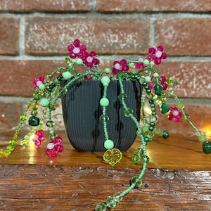 Könnte beinhalten: Ein dekoratives Blumenarrangement aus Perlen in einem schwarzen Topf. Das Arrangement zeigt rosa Blüten, grüne Perlen und hängende Akzente. Der Topf steht auf einer Holzoberfläche vor einem Ziegelhintergrund. Das Perlendesign verleiht der Blumenausstellung eine einzigartige Note.