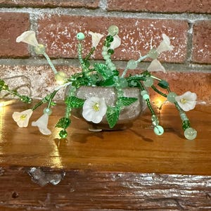 Pode incluir: Um arranjo floral decorativo em um pequeno vaso castanho claro texturizado. O arranjo apresenta flores brancas translúcidas, folhas verdes e contas verdes. O fundo é uma parede de tijolos e uma superfície de madeira.