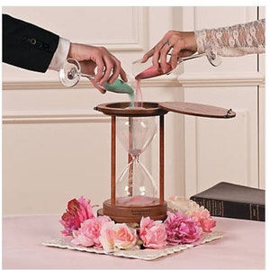 Pu&ograve; includere: Una clessidra di legno con un coperchio, utilizzata per una cerimonia di sabbia. Due persone versano sabbia colorata da contenitori di vetro nella clessidra. La clessidra &egrave; decorata con fiori rosa.
