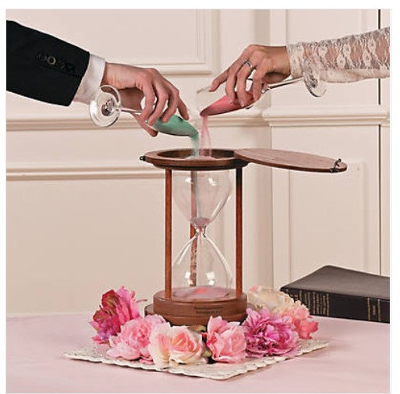 Pu&ograve; includere: Una clessidra di legno con un coperchio, utilizzata per una cerimonia di sabbia. Due persone versano sabbia colorata da contenitori di vetro nella clessidra. La clessidra &egrave; decorata con fiori rosa.