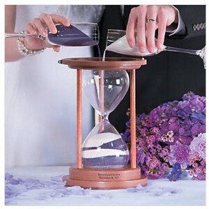 Pu&ograve; includere: Una clessidra di legno con due scomparti, uno riempito di sabbia viola e l'altro di sabbia bianca. La clessidra viene utilizzata durante una cerimonia di matrimonio, con due persone che versano sabbia da contenitori separati nella clessidra.