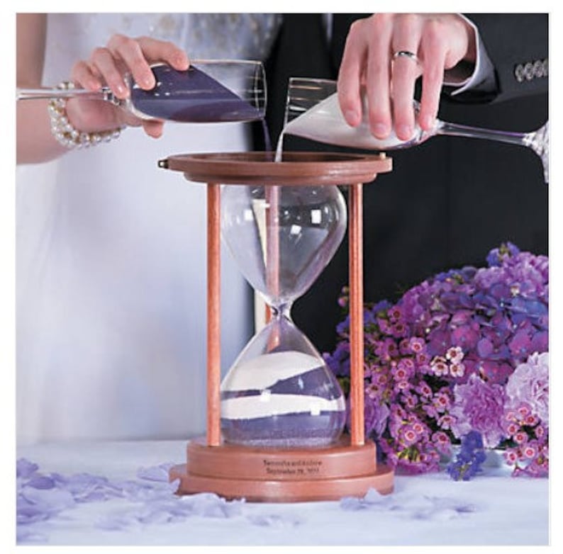 Pu&ograve; includere: Una clessidra di legno con due scomparti, uno riempito di sabbia viola e l'altro di sabbia bianca. La clessidra viene utilizzata durante una cerimonia di matrimonio, con due persone che versano sabbia da contenitori separati nella clessidra.