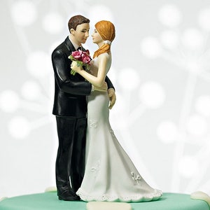 May include: A wedding cake topper featuring a bride and groom figurine. The bride is wearing a white gown with lace detailing and a long veil. The groom is wearing a black tuxedo with a white shirt and a black bow tie. The couple is holding a bouquet of pink roses.