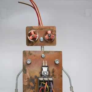 May include: A robot sculpture constructed from electronic components, featuring a rectangular copper torso with a central terminal block, copper wire limbs, a circuit board base, and a head with two copper coils and protruding wires.