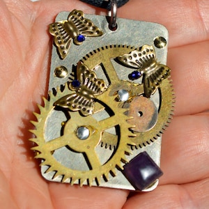 Puede incluir: Un colgante rectangular de metal en un cordón negro que presenta tres dijes de mariposa en tono latón con pequeños diamantes de imitación azules, dos engranajes de reloj superpuestos y una piedra preciosa cuadrada de color púrpura.