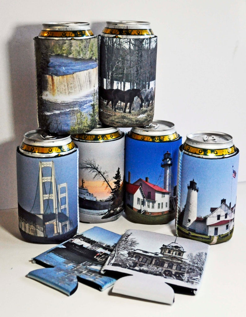 May include: A set of seven colorful can coolers with various images printed on them. The images include a waterfall, a forest, a bridge, a lighthouse, a sunset, and a building. The can coolers are made of foam and are designed to keep drinks cold.