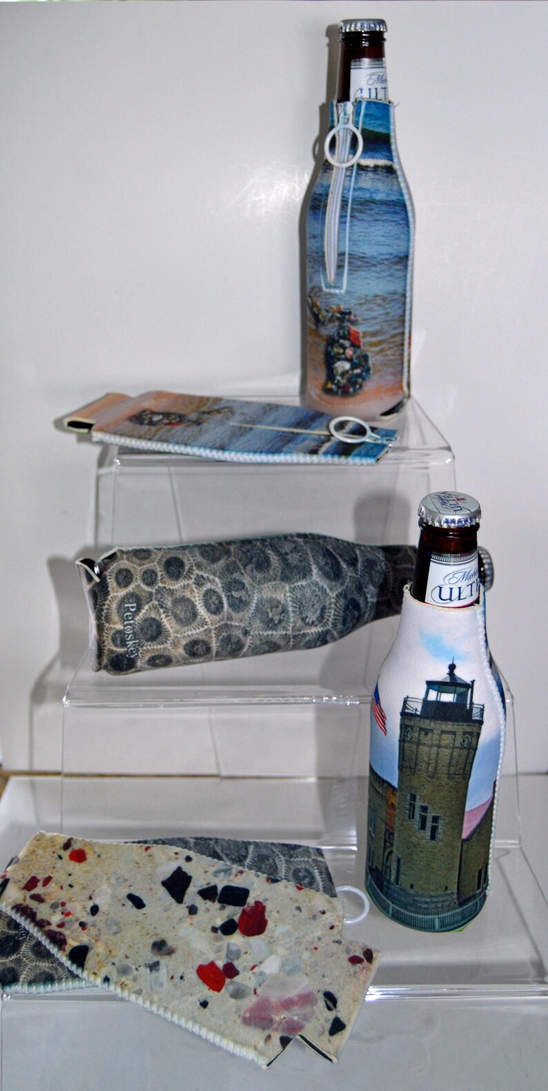 May include: Three different bottle coolers with zipper closures. The first cooler is blue and white with a picture of a boat. The second cooler is brown and white with a pattern of fossilized stones. The third cooler is white with a picture of a lighthouse. All three coolers have a zipper closure.