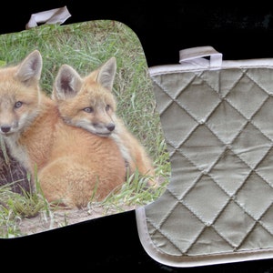 May include: A square pot holder featuring a print of two fox cubs nestled in grass. The reverse side is a quilted pattern in a muted green color with a white trim and a hanging loop. Kitchen and home decor.
