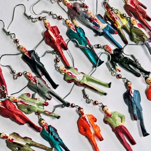 May include: A collection of zombie-themed earrings featuring miniature figures in various poses and clothing. The figures are painted in shades of red, green, blue, orange, and black, with blood splatters for a gruesome effect.