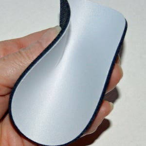 May include: A white fabric patch with a black trim, folded in half and held in a hand. The fabric is smooth and appears to be a type of foam.