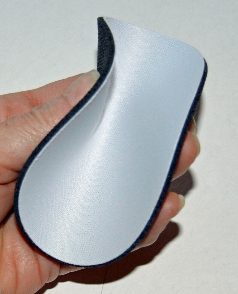 May include: A white fabric patch with a black trim, folded in half and held in a hand. The fabric is smooth and appears to be a type of foam.