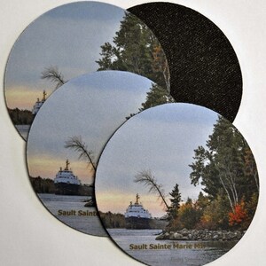 May include: Four round coasters with a scenic image of a ship on a river with trees and a cloudy sky. The coasters have a black backing and the text "Saulte Sainte Marie MI" printed on the bottom.