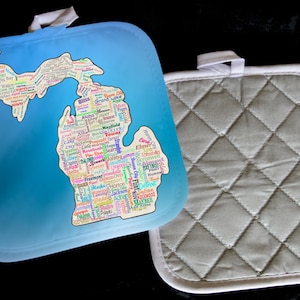 May include: A blue and white pot holder with a map of Michigan made of city names. The map is surrounded by a blue background. The pot holder is quilted and has a white border.