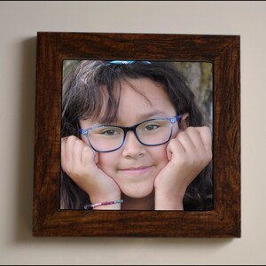 May include: A young person with brown hair wearing blue-rimmed glasses and a blue bracelet, smiling with their hands on their cheeks.