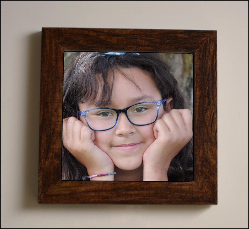 May include: A young person with brown hair wearing blue-rimmed glasses and a blue bracelet, smiling with their hands on their cheeks.