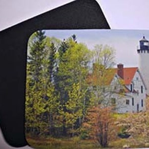 May include: A white lighthouse with a red roof stands tall against a backdrop of green trees and a blue sky. The lighthouse is surrounded by rocks and bushes.