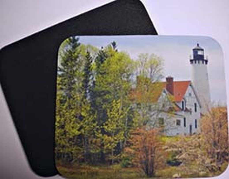 May include: A white lighthouse with a red roof stands tall against a backdrop of green trees and a blue sky. The lighthouse is surrounded by rocks and bushes.