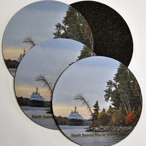 May include: Four round coasters with a scenic image of a ship on a river with trees and a cloudy sky. The coasters have a black backing and the text "Saulte Sainte Marie MI" printed on the bottom.