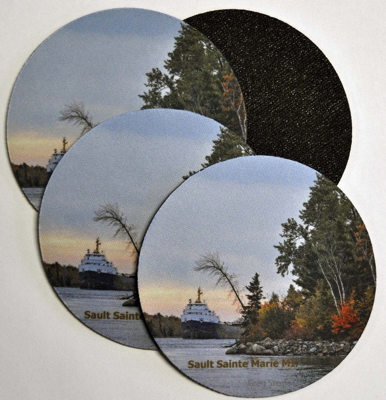 May include: Four round coasters with a scenic image of a ship on a river with trees and a cloudy sky. The coasters have a black backing and the text "Saulte Sainte Marie MI" printed on the bottom.