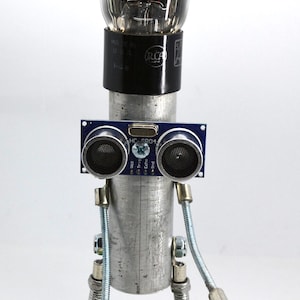 May include: A robot made of recycled materials, including a glass vacuum tube, a circuit board, and metal parts. The robot has a blue and black sensor attached to the front, and a silver metal body. The robot is standing on a green circuit board.