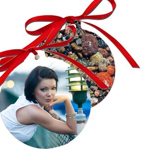 May include: A round Christmas ornament with a red ribbon. The ornament features a photo of a woman with short black hair, wearing a white top, leaning on a railing. The background is a blurry image of a street scene with a streetlight.
